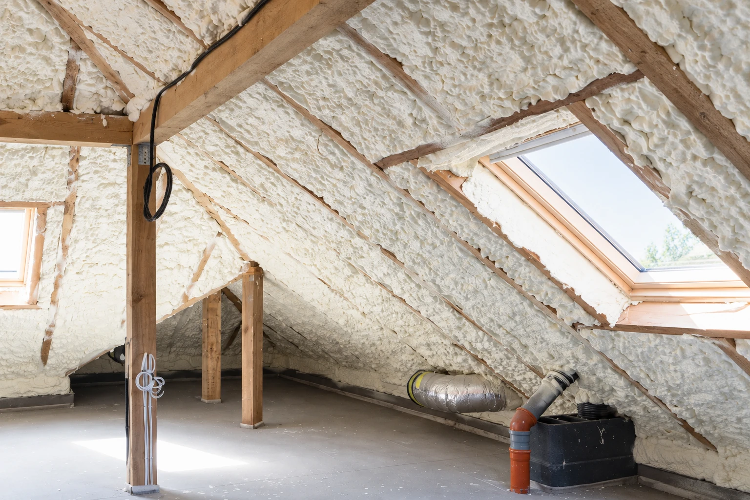 Attic with white spray foam covering the sloped roof and rafters, with a skylight, support posts, and ductwork.