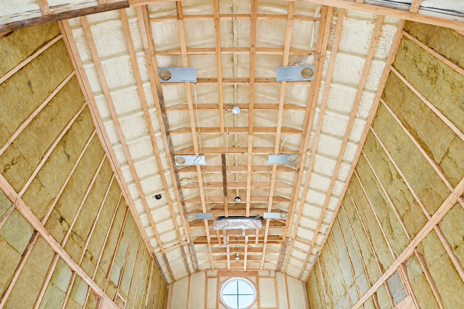 Attic roof and walls are lined with pale foam insulation between wooden framing, with a round window at the far end.