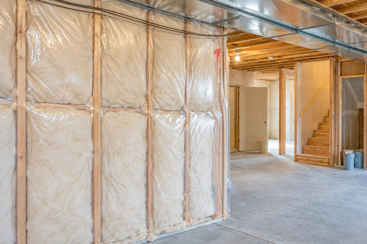 Basement wall with fiberglass insulation between wooden studs, covered by clear plastic sheeting beside an open unfinished room.