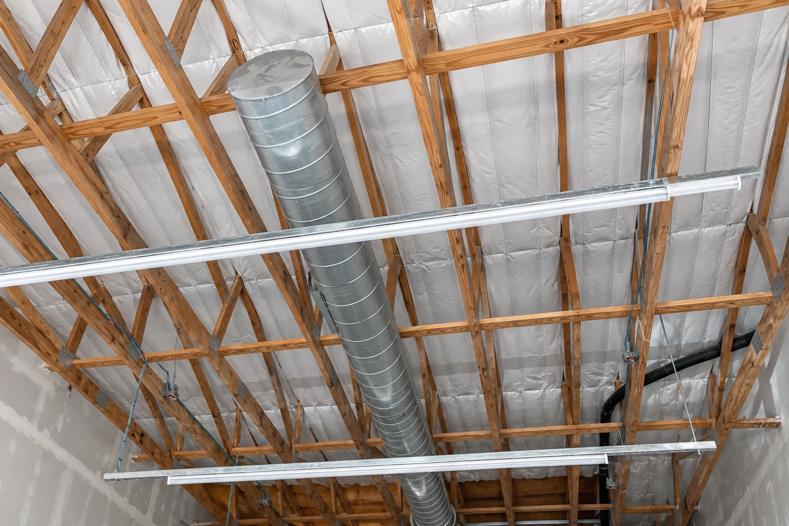 Reflective insulation lines a commercial building’s exposed timber roof structure around a large metal ventilation duct and light fixtures.
