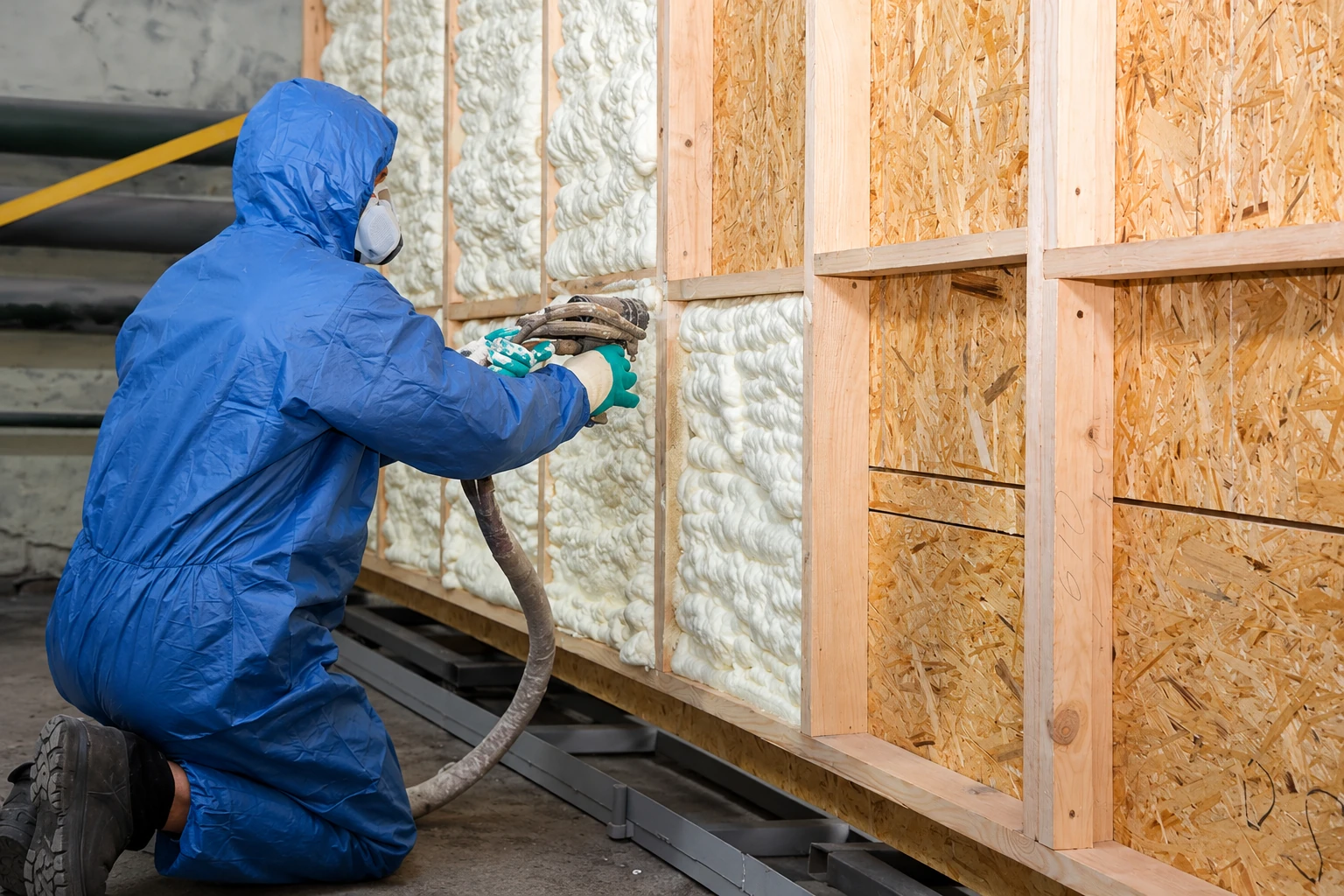 Worker in blue protective suit sprays foam insulation into framed wall cavities at a construction site.