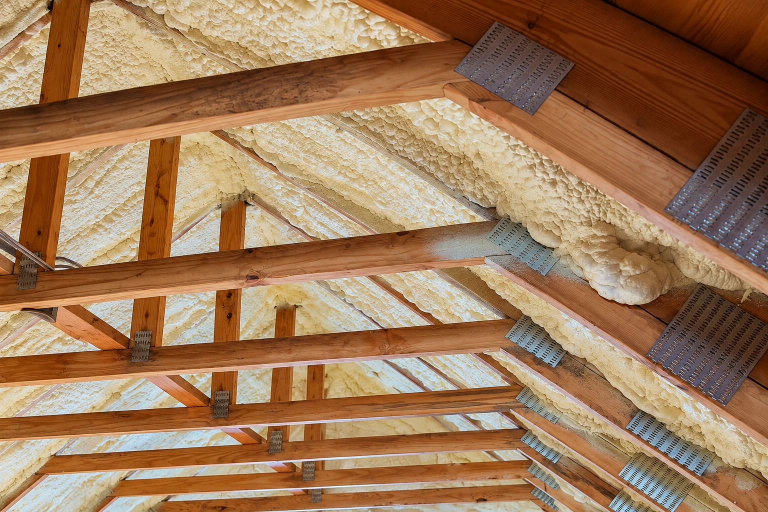 Spray foam insulation coats the underside of a wooden roof, filling the spaces between exposed rafters and trusses.